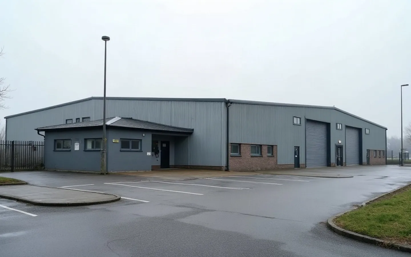 Een laag bedrijfspand met een kantoordeel en een hal met gesloten roldeuren, gezien vanaf een leeg parkeerterrein in het ochtendlicht.
